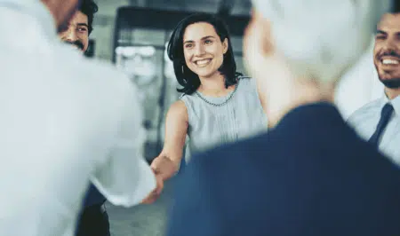 A smiling woman in business attire shakes hands with someone in an office setting, surrounded by colleagues who are also smiling.