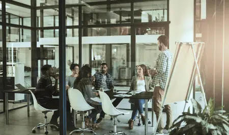 A group of six people sit around a table in a modern office meeting room, listening to a man standing next to a blank flip chart, with large windows in the background letting in natural light.