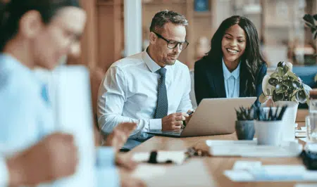 Three professionals sit at a desk in an office. A man and a woman, both in business attire, focus on a laptop and smile, while another person in the foreground is blurred. There are office supplies and a plant on the desk.