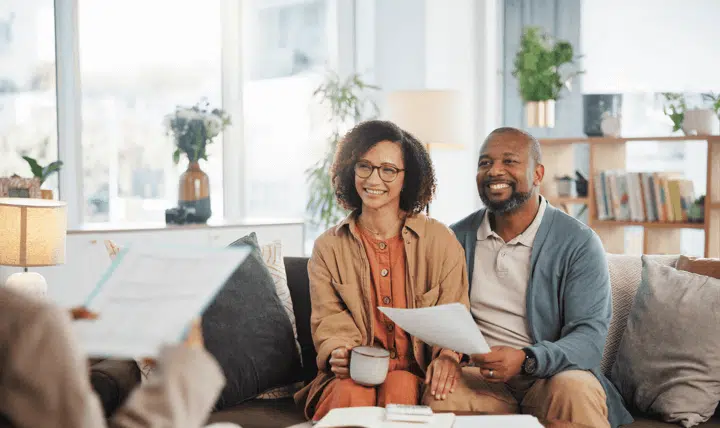A smiling couple sits together on a couch, holding papers and a coffee mug, while talking to a person off-camera in a bright, cozy living room filled with plants and books.
