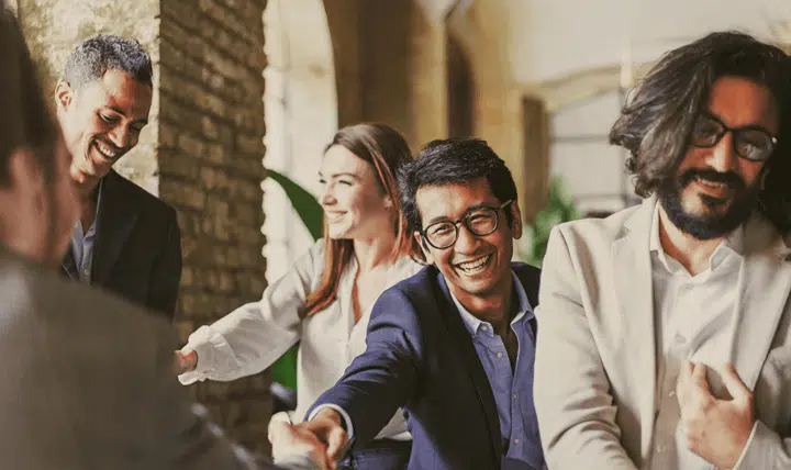 A group of four professionally dressed people laugh and shake hands in a bright, modern office space, appearing happy and engaged in conversation.