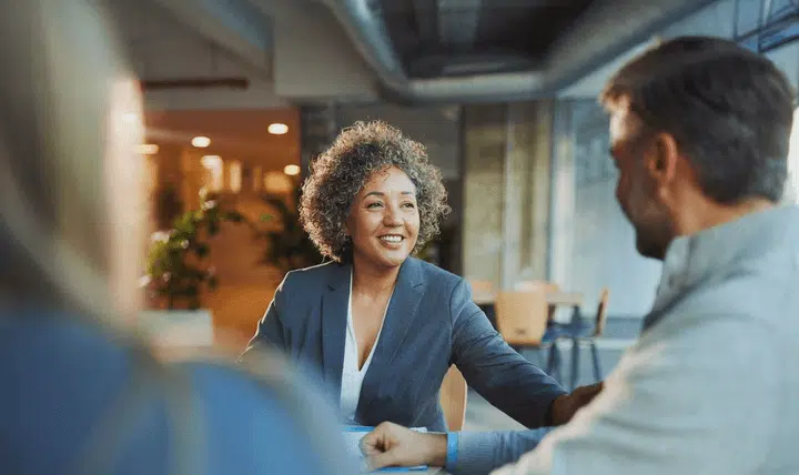 A woman in a gray blazer smiles and talks with two people sitting across from her in a modern, brightly lit office space with large windows and plants in the background.