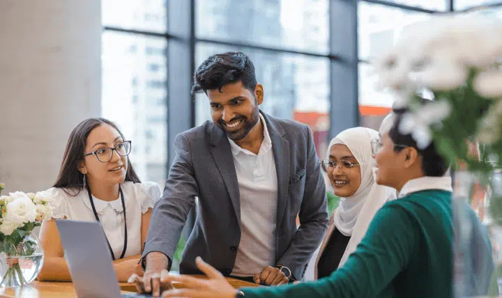 Four professionally dressed people gather around a laptop in a modern office, smiling and engaged in discussion. Large windows and flowers are visible in the background, creating a bright, collaborative atmosphere.