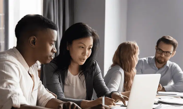 Four people sit at a table working together. Two are focused on a laptop in the foreground, while two others in the background have a discussion. The setting appears to be a modern office or meeting room.