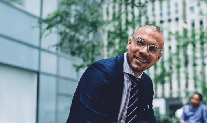 A man wearing glasses, a navy suit, and a striped tie smiles while sitting outdoors in an urban setting with greenery and modern buildings in the background.