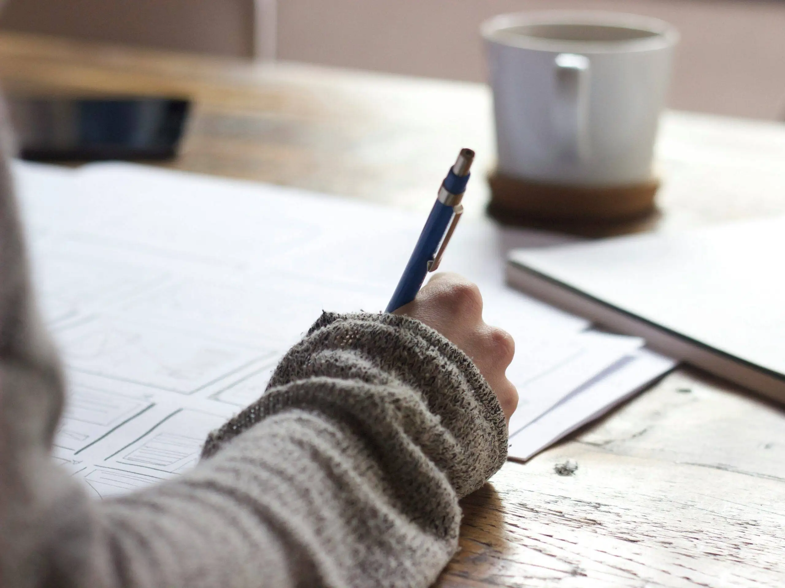 A person wearing a gray sweater writes on paper at a wooden desk, with a pen in hand. A white mug and a phone are also visible on the table.