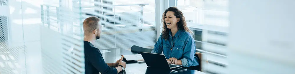 Two people sit across from each other at a table in a modern office, smiling and talking. One has a laptop open in front of them. Glass walls and bright natural light fill the space.