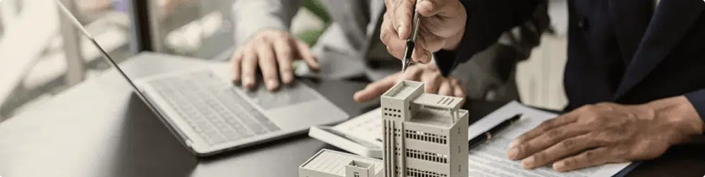 Two people sit at a desk with a laptop and documents. One person holds a key above a model of buildings, suggesting a real estate or property transaction.