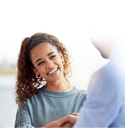 A smiling woman with curly hair shakes hands with another person, both appearing happy, in a bright indoor setting.